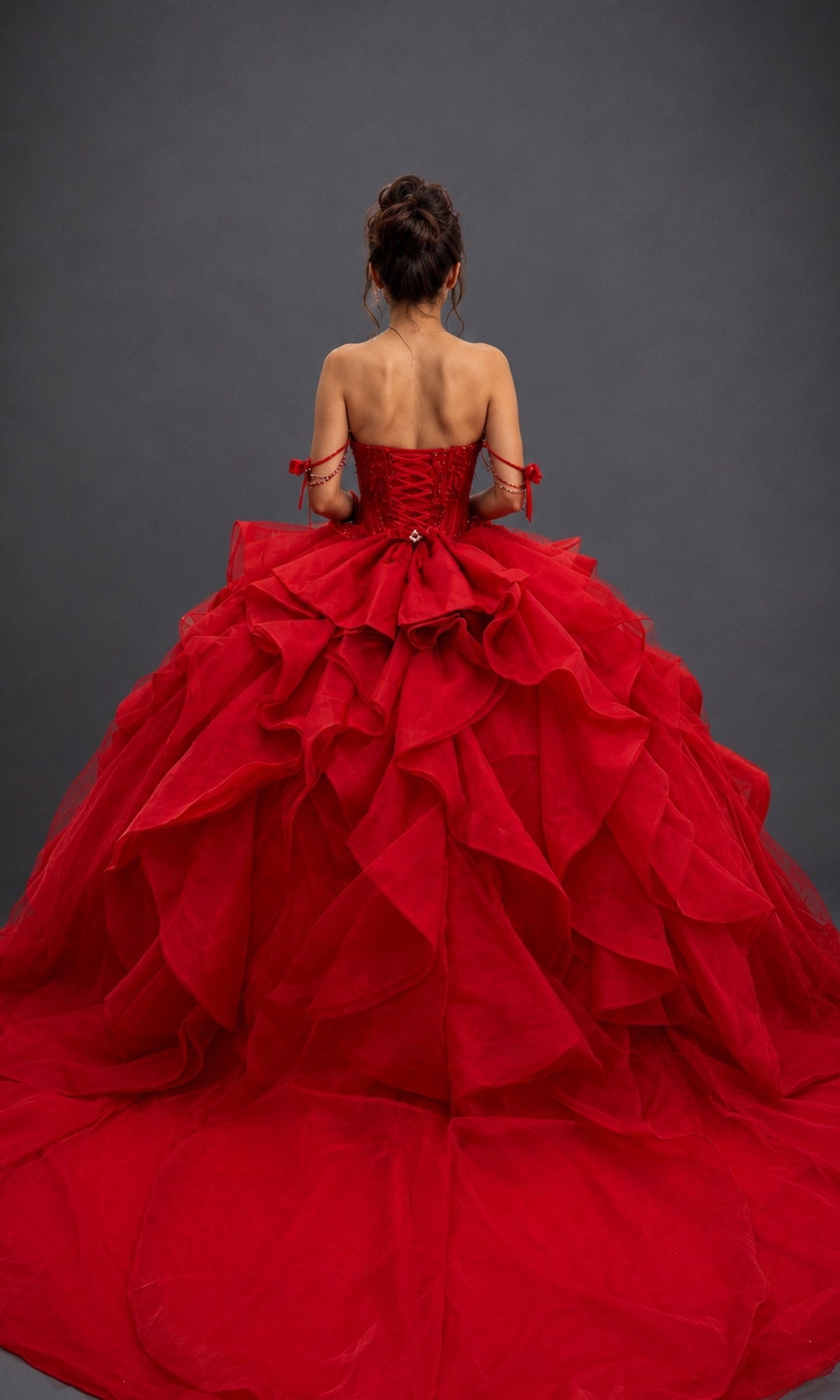 Wearing the Quinceanera Dress 5197 By Dancing Queen, a woman stands with her back to the camera, showcasing the gown’s strapless red beaded corset bodice and voluminous ruffled tulle skirt against a plain dark background.