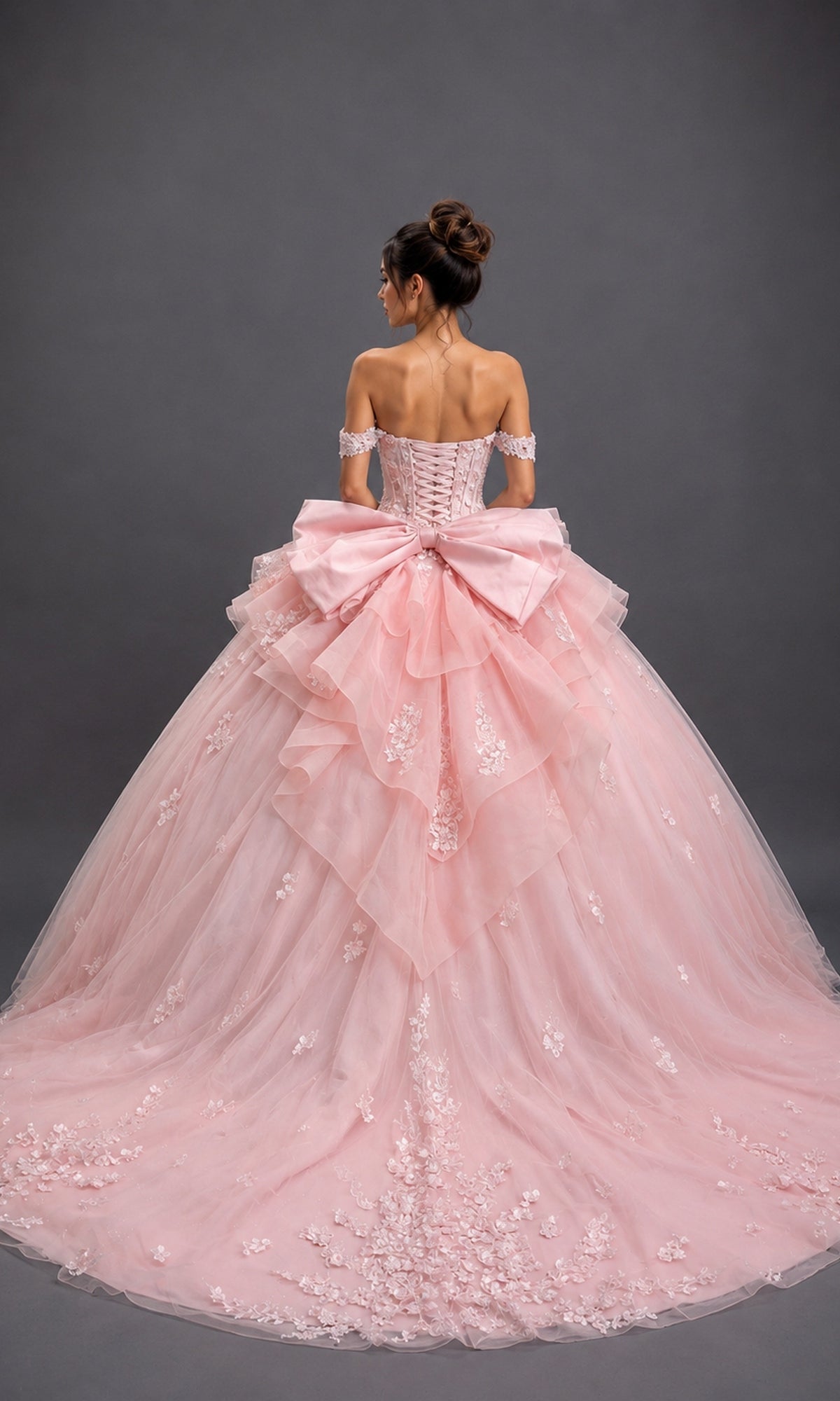 A woman stands facing away against a plain gray background, wearing the Quinceanera Dress 5200 By Dancing Queen—an off-the-shoulder pastel pink ballgown featuring a sweetheart neckline, delicate floral details, and a large bow.