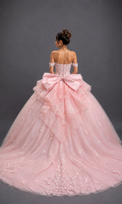 A woman stands facing away against a plain gray background, wearing the Quinceanera Dress 5200 By Dancing Queen—an off-the-shoulder pastel pink ballgown featuring a sweetheart neckline, delicate floral details, and a large bow.