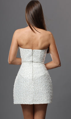 A woman facing away wears the Jovani 52095 short homecoming dress, a white strapless mini dress crafted from textured fabric and adorned with delicate hand-beaded pearl embellishments, set against a plain gray background.