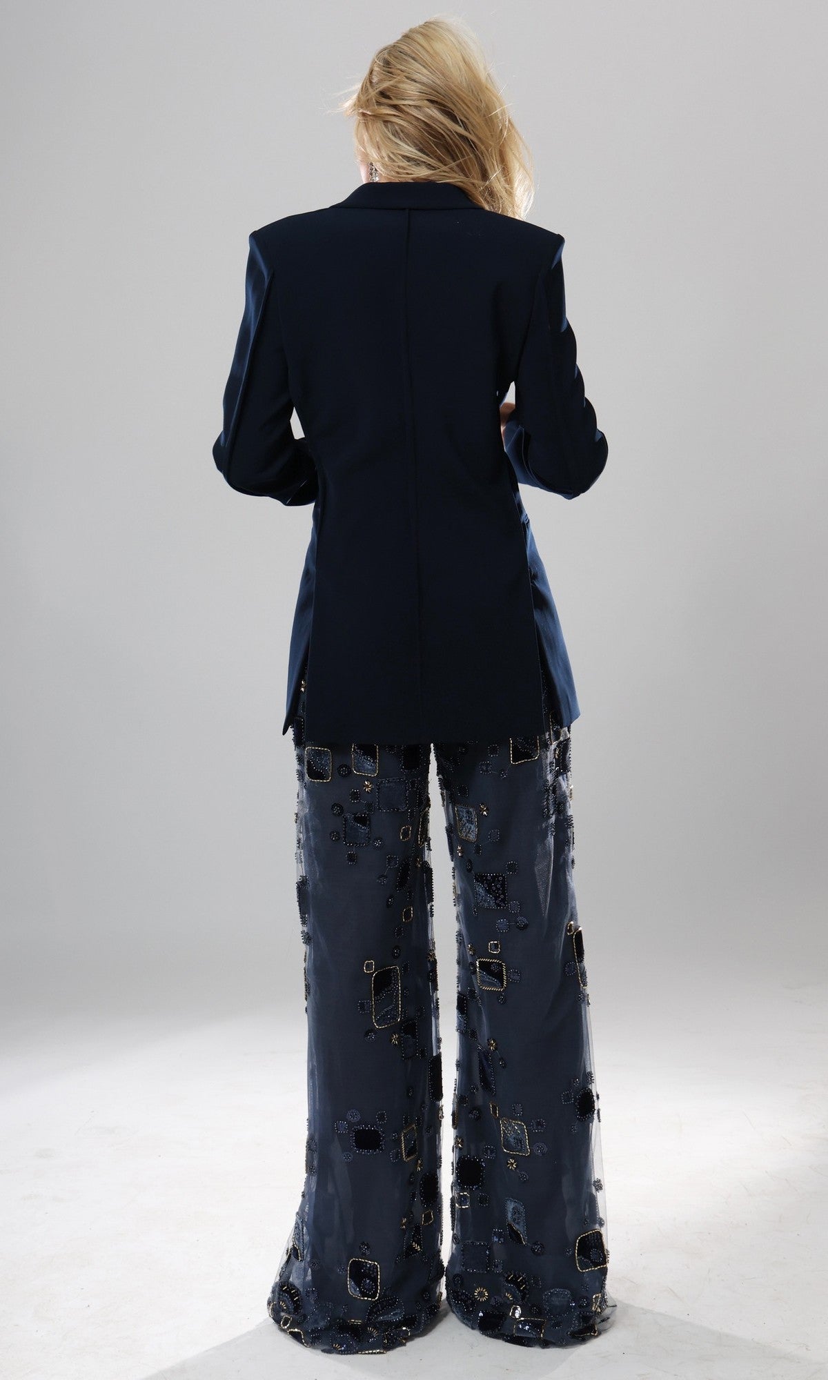 A person stands with their back to the camera, wearing the Long Formal Jumpsuit: Jovani 52096—a dark designer one-piece featuring wide-leg pants adorned with square and circle patterns—set against a plain, light background.
