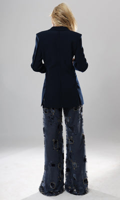 A person stands with their back to the camera, wearing the Long Formal Jumpsuit: Jovani 52096—a dark designer one-piece featuring wide-leg pants adorned with square and circle patterns—set against a plain, light background.