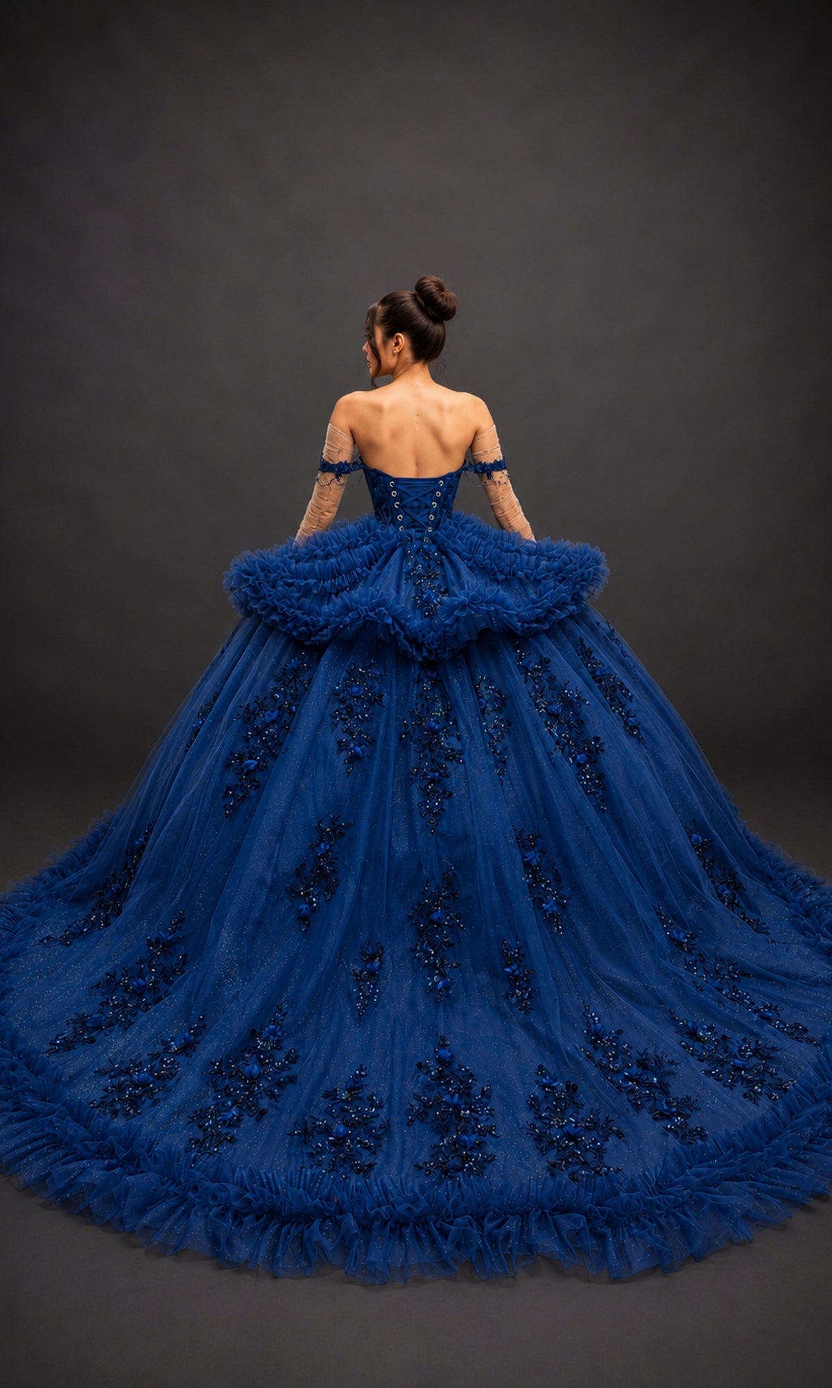A woman stands facing away against a dark background, wearing the Quinceanera Dress 5215 by Dancing Queen—a navy-blue gown featuring an off-the-shoulder sweetheart neckline, adorned with lace and floral embellishments.