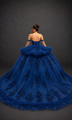 A woman stands facing away against a dark background, wearing the Quinceanera Dress 5215 by Dancing Queen—a navy-blue gown featuring an off-the-shoulder sweetheart neckline, adorned with lace and floral embellishments.
