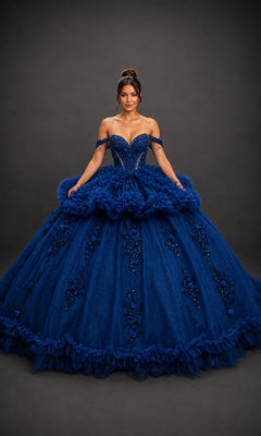 A woman stands against a dark background wearing the Quinceanera Dress 5215 by Dancing Queen in navy. The dramatic gown features ruffles, sequins, and floral embellishments. Her hair is styled up with a headpiece that beautifully complements the off-the-shoulder sweetheart neckline of her dazzling ensemble.