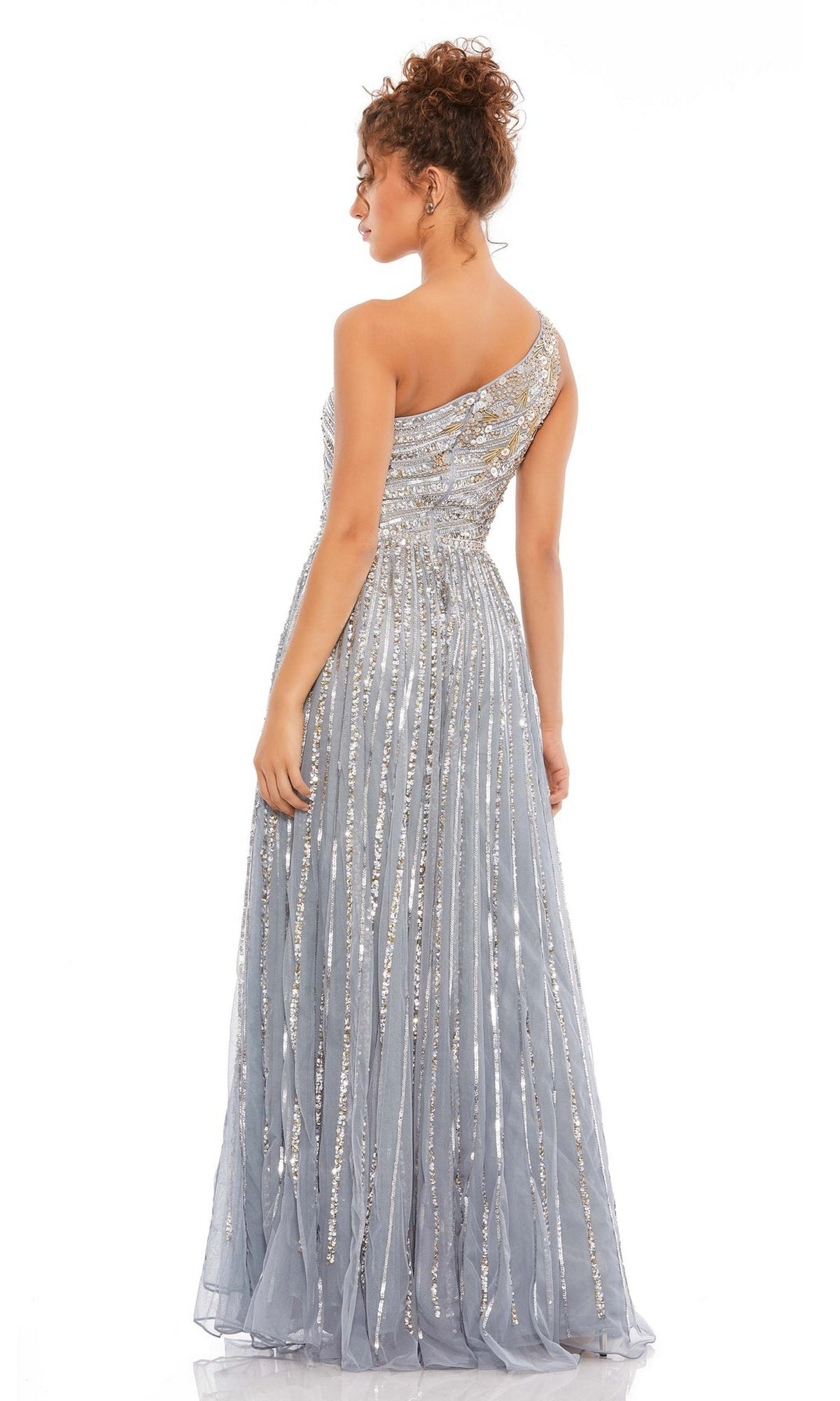 A woman stands facing away, wearing the Mac Duggal 5220 One-Shoulder Beaded Formal Dress in light blue with silver sequin embellishments and floor-length design.