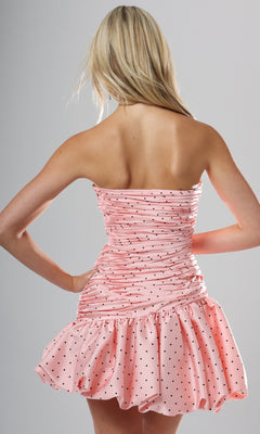 A woman with long blonde hair models the Jovani 52226 short homecoming dress, showcasing its strapless pink polka-dot design, ruched bodice, and short a-line bubble skirt as she is seen from the back against a plain gray background.