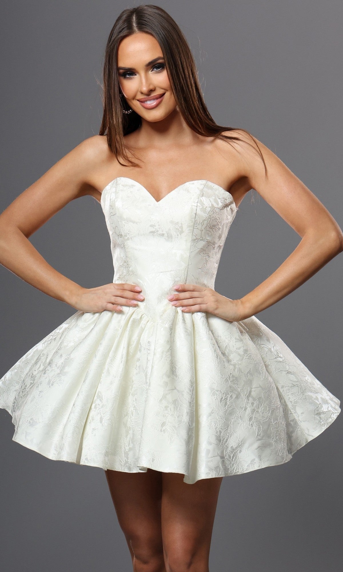 A woman stands against a gray background, smiling with her hands on her waist, wearing the Jovani 52228 short homecoming dress in white—an A-line mini style with a strapless neckline and flared skirt.