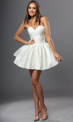 A woman wearing the Short Homecoming Dress: Jovani 52228—a strapless, white, a-line mini dress with a flared skirt—paired with clear high heels, stands and smiles against a gray background.