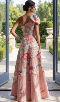 Wearing the Jovani 52377 Long Formal Dress in pink, a woman stands with her back to the camera in a sunlit room with open double doors, looking out onto a garden. The one-shoulder floral brocade gown catches the light, accentuating its delicate details and elegant silhouette.