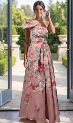 A woman wearing the Jovani 52377 Long Formal Dress in Pink Multi stands indoors by open glass doors, with a lush garden visible in the background. The elegant gown features delicate floral brocade detailing, adding a touch of sophistication to the scene.