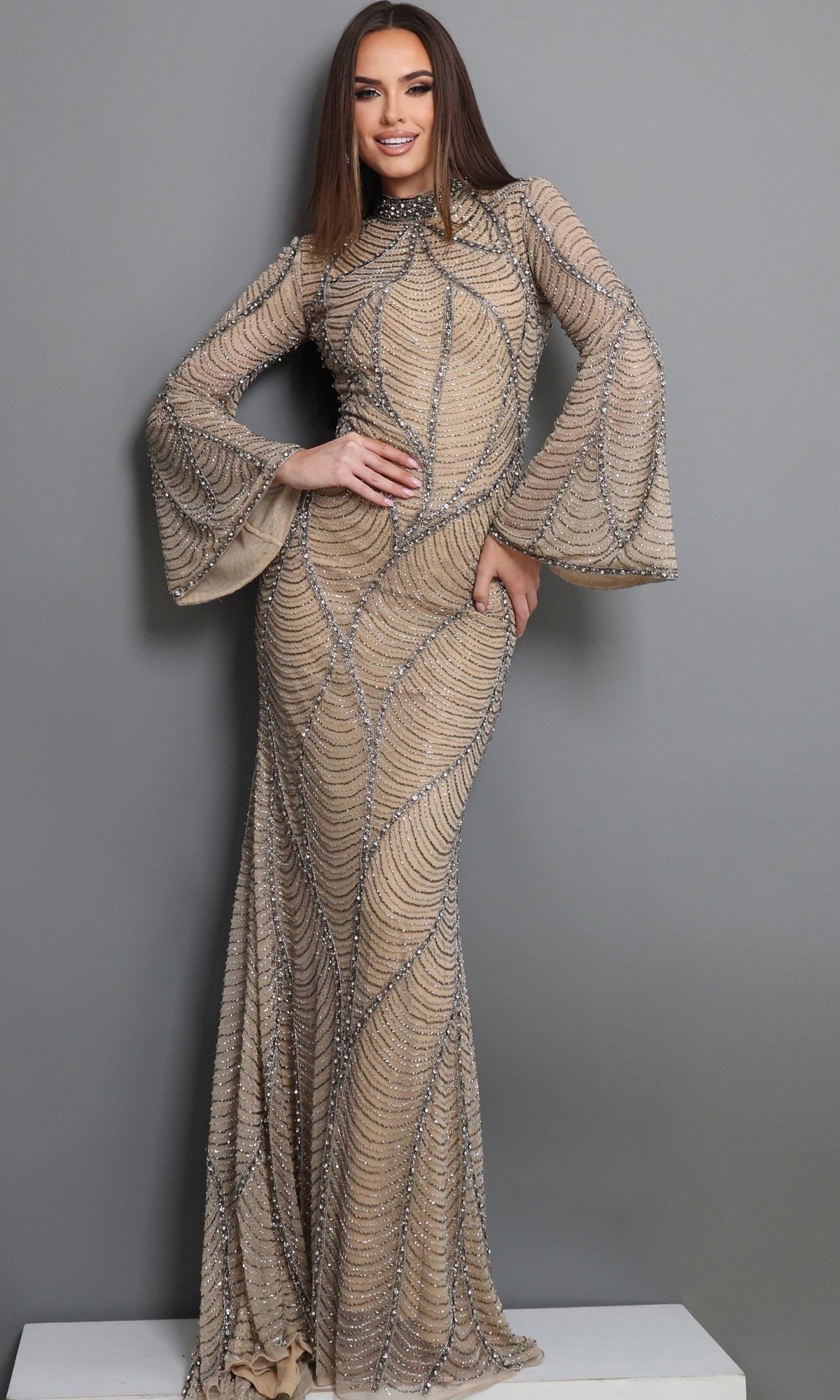 A woman with straight brown hair wears the Jovani 52447 Long Formal Dress in Nude/Silver, featuring silver beaded details and elegant flared sleeves, as she stands against a gray background—a sophisticated evening gown ideal for the mother of the bride.