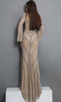 A woman with long brown hair stands on a white block, facing away, modeling the Long Formal Dress: Jovani 52447—a beige, floor-length modest gown with long sleeves and intricate patterns—against a plain gray background.