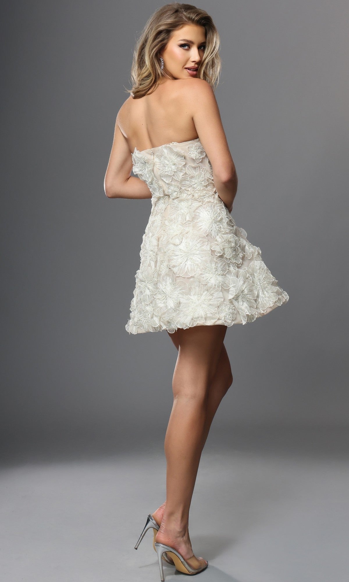 A woman faces away, wearing the Jovani 52558 short homecoming dress—a strapless, fit-and-flare mini dress crafted from textured fabric with delicate 3D petal appliques—paired with high heels and standing against a plain gray background.