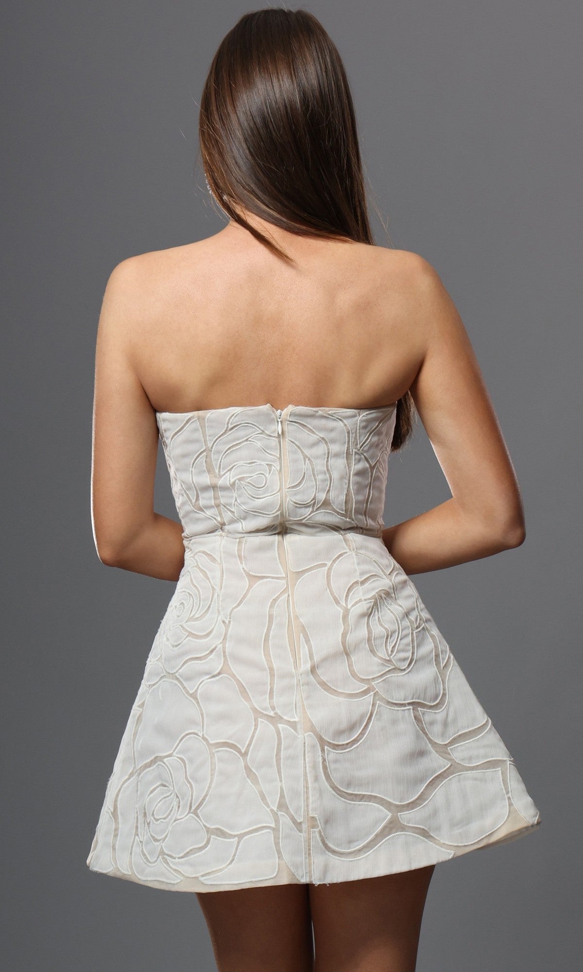 A woman with long brown hair is shown from the back against a gray background, wearing the Jovani 52560 short homecoming dress—a white strapless fit-and-flare silhouette adorned with a floral pattern.