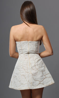 A woman with long brown hair is shown from the back against a gray background, wearing the Jovani 52560 short homecoming dress—a white strapless fit-and-flare silhouette adorned with a floral pattern.