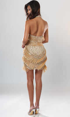 A woman stands with her back to the camera against a plain light background, wearing the Jovani 52605 short homecoming dress—a halter mini dress adorned with gold beaded fringe—paired with beige high heels.