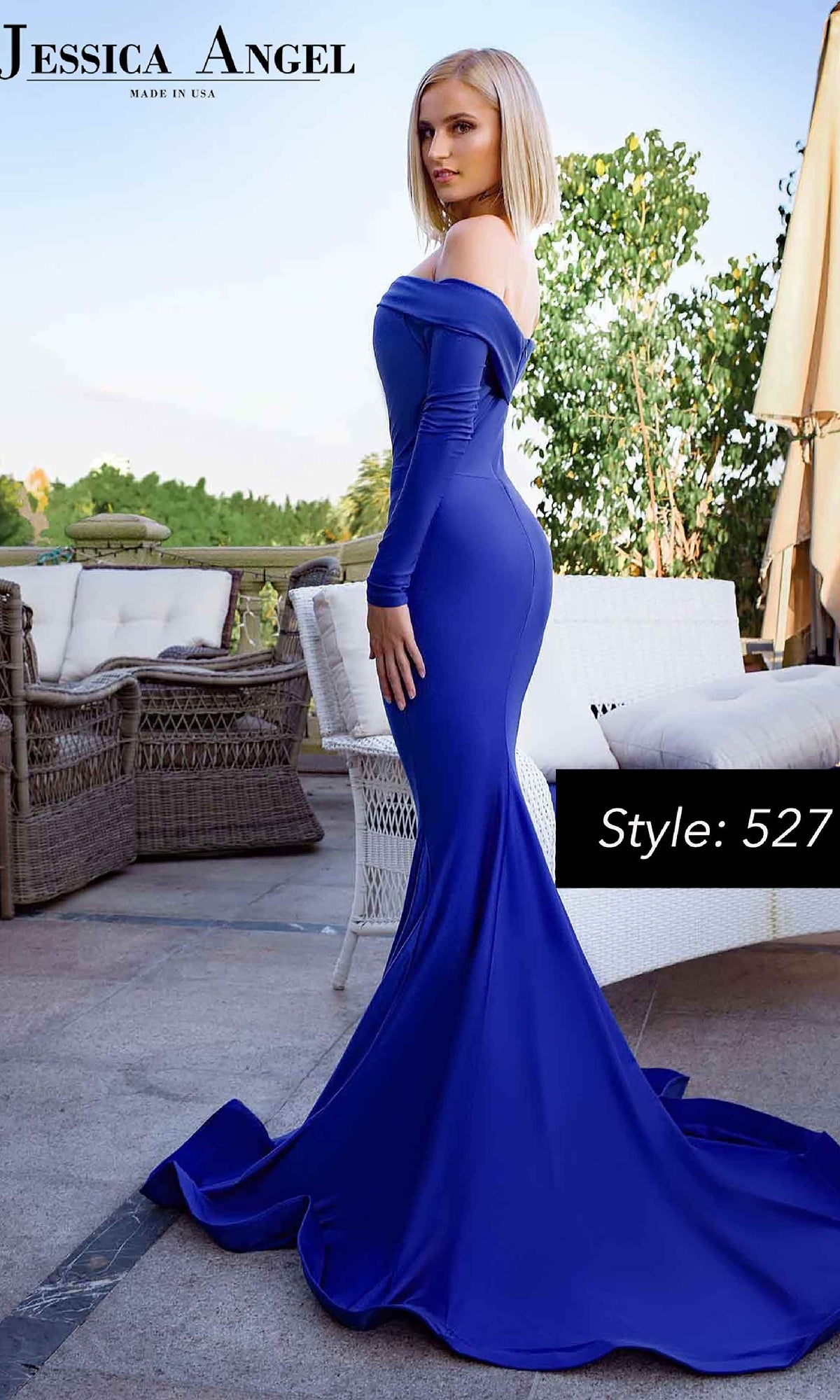 A woman in a fitted, long sleeve Jessica Angel 527 prom gown with a mermaid skirt and flowing train stands on a patio. The off-the-shoulder, royal blue dress is elegant and stylish—perfect for your next formal event.