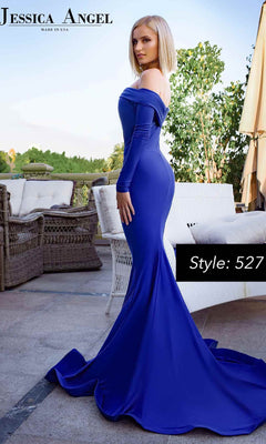 A woman in a fitted, long sleeve Jessica Angel 527 prom gown with a mermaid skirt and flowing train stands on a patio. The off-the-shoulder, royal blue dress is elegant and stylish—perfect for your next formal event.