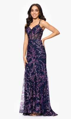 Woman wearing the Blondie Nites 5273BN Navy Blue Print Prom Dress, featuring thin straps, sheer panels, and pink floral patterns, stands and smiles with one hand on her hip.