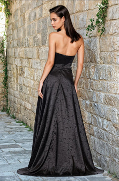 A woman with straight dark hair models the Strapless Black Prom Gown: Blondie Nites 5301BN, standing with her back to the camera against a stone wall covered in green vines.