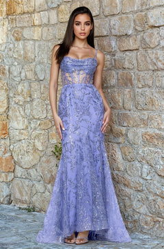 A woman stands before a stone wall wearing the Long Periwinkle Prom Dress Blondie Nites 5315BN, a fitted mermaid gown with sequin accents and sheer corset panels.