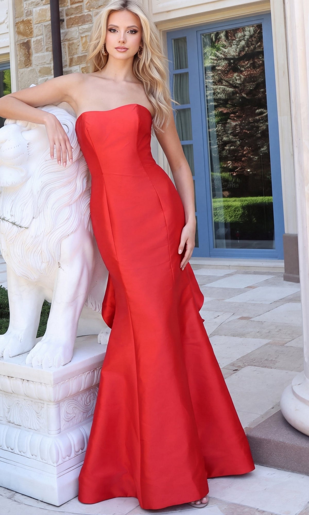 A woman in a red Jovani 53235 strapless ruffle trumpet gown leaning against a white stone lion statue.