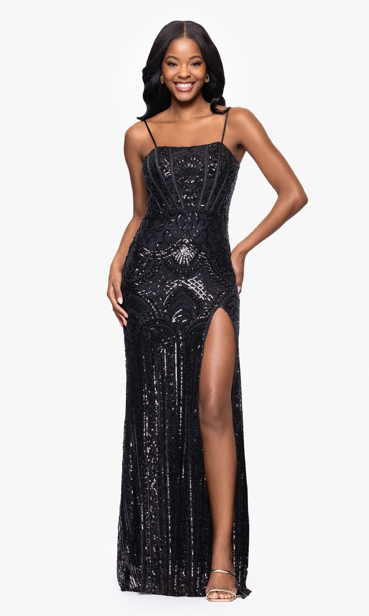 A woman smiles while wearing the Blondie Nites 5327BN long black sequin prom dress with thin straps, intricate patterns, and a high slit, standing against a plain white background.
