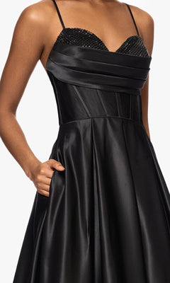 A woman wearing the Long Black A-Line Prom Dress (Blondie Nites 5333BN) with thin straps, a beaded bodice, sleeveless design, and her right hand in a pocket.