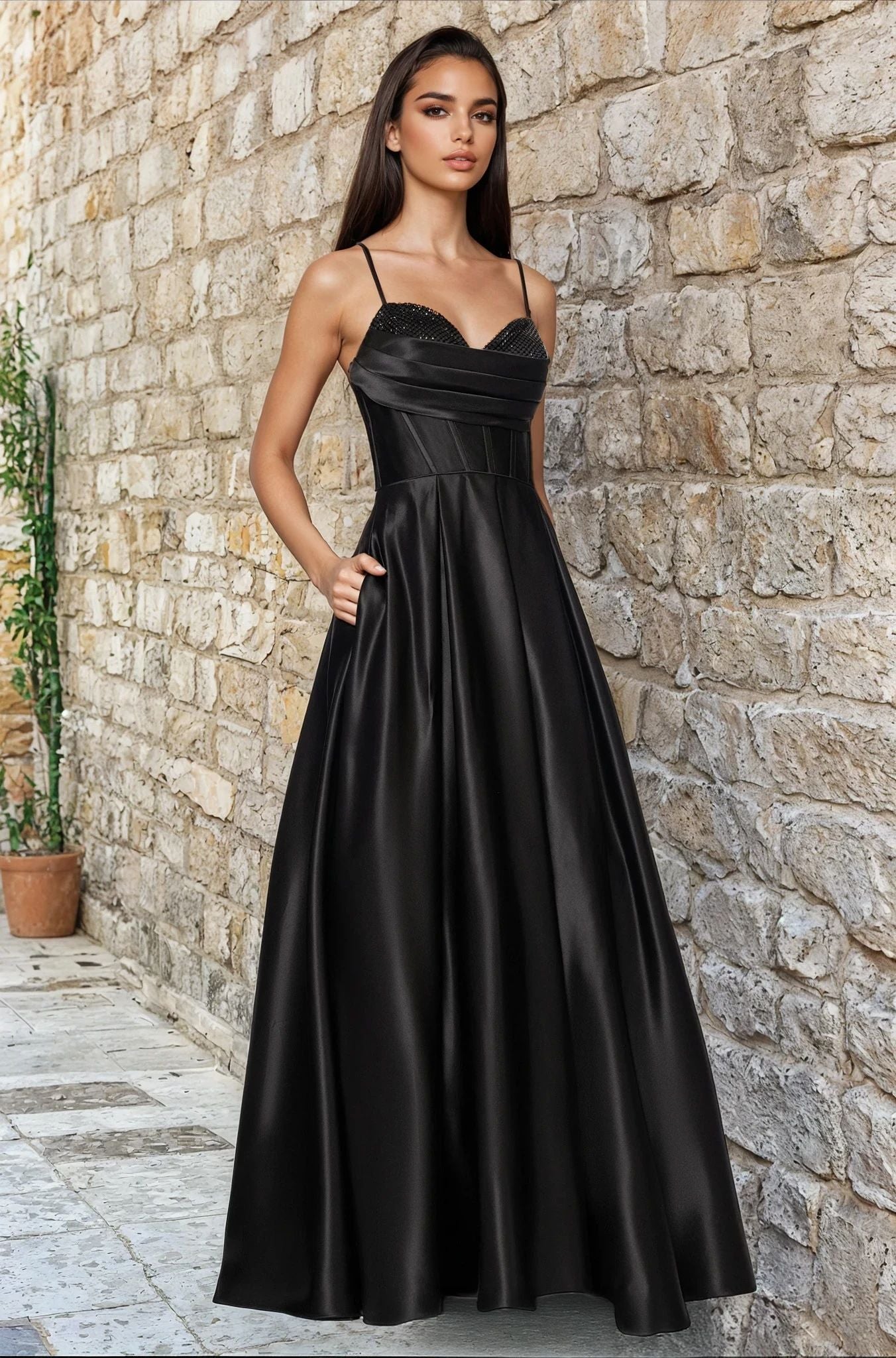 A woman stands gracefully against a stone wall, wearing the Blondie Nites 5333BN—a long black A-line prom dress with thin straps—styled for a flattering silhouette.