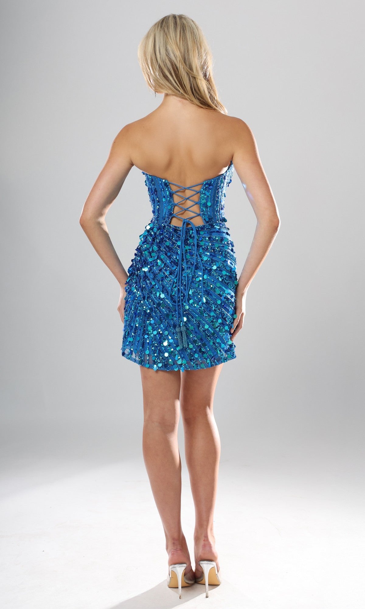 A woman stands with her back to the camera, showcasing the Jovani 53770 short homecoming dress—a blue strapless sequin dress featuring a corset bodice with lace-up open back detailing—paired with high heels, against a plain background.