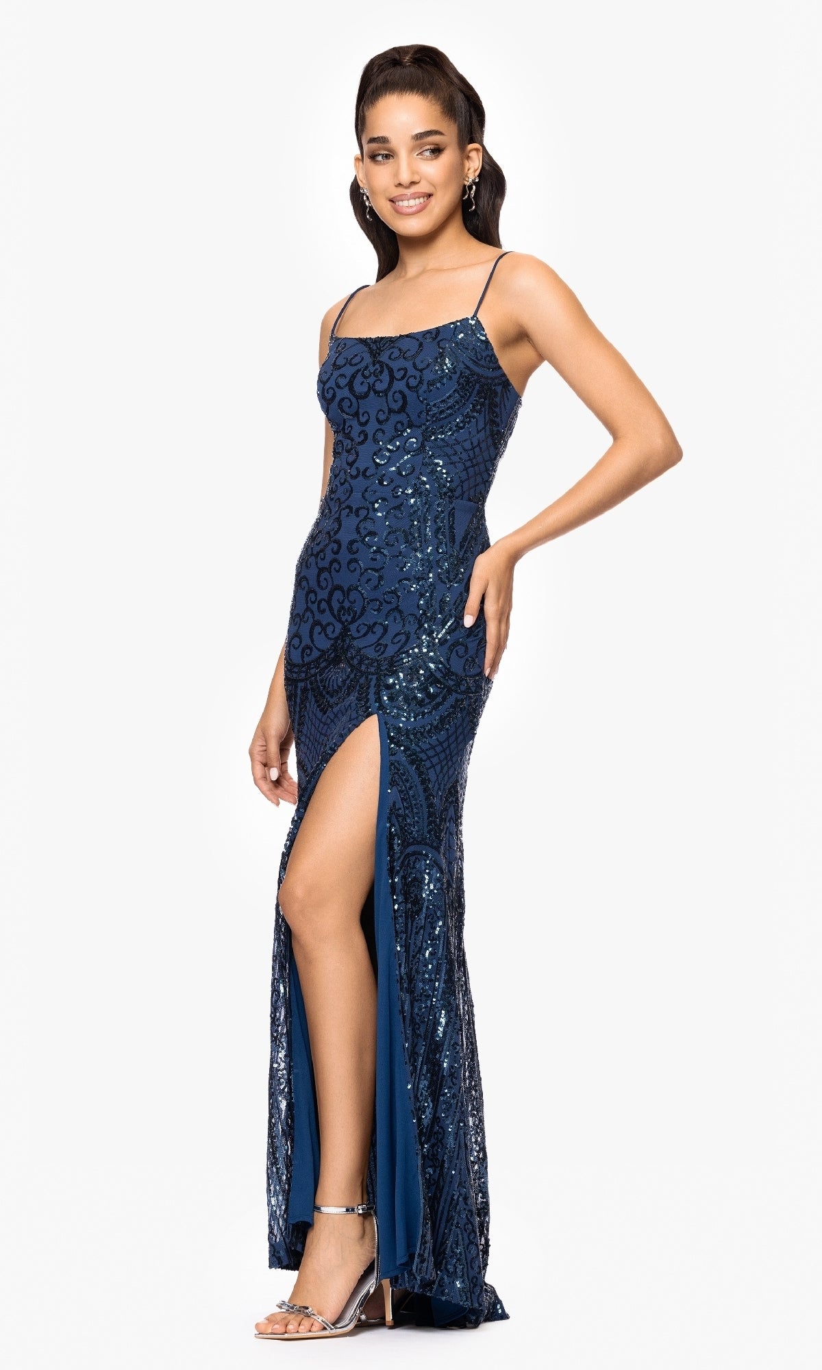 A woman in the Blondie Nites 5377BN long sequin-print prom dress, featuring thin straps and a high thigh slit, stands against a white background styled with silver heels and earrings.
