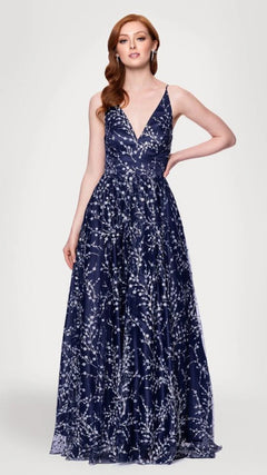 A woman stands with hand on hip in the Blondie Nites 5386BN long prom dress, featuring a sleeveless, deep V-neck navy/peri design with a white floral pattern and an elegant A-line skirt, against a plain light gray background.