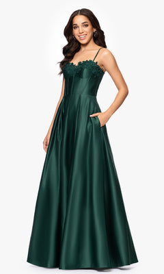 A woman stands smiling against a plain white background, wearing the Classic Long Prom Dress: Blondie Nites 5387BN—a dark green satin gown with embroidered-lace corset trim, thin straps, and pockets.