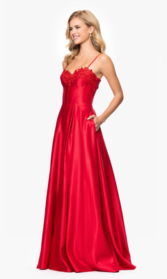 A woman smiles as she stands with one hand in her pocket, wearing the Classic Long Prom Dress: Blondie Nites 5387BN, which features a bright red satin skirt, thin straps, and an embroidered lace corset bodice.
