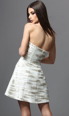A woman stands with her back to the camera against a plain gray background, wearing the Jovani 53953 short homecoming dress—a strapless white design featuring metallic gold patterns.