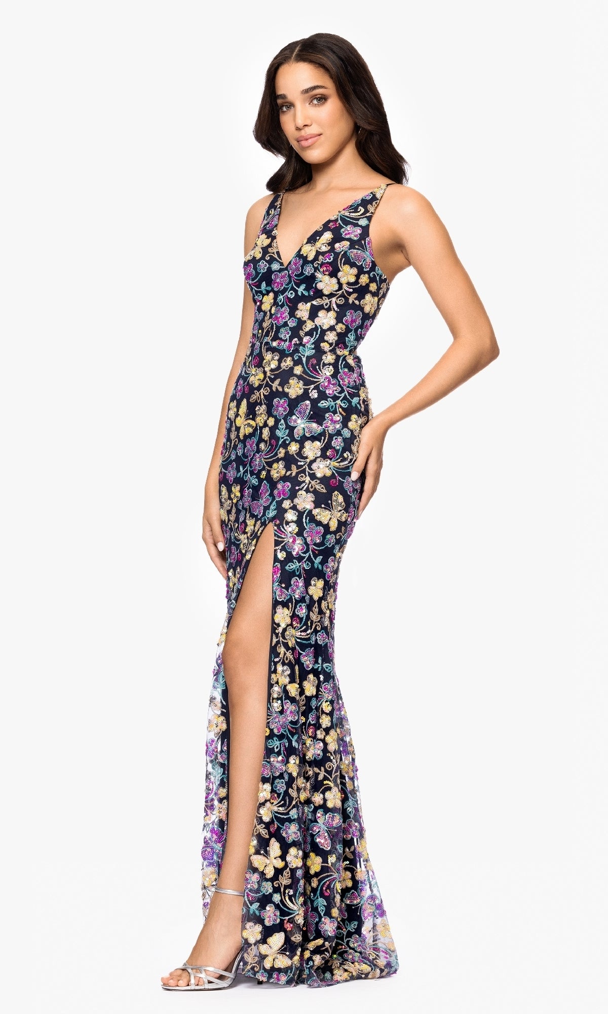 Woman wears the Sequin Butterfly Prom Gown: Blondie Nites 5402BN, a sleeveless, floor-length black dress with a colorful butterfly sequin pattern and high slit, paired with silver heels against a white background.