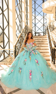 A woman wears the Quinceanera Dress 54200 By Amarra, a large light blue tulle ball gown with floral embroidery, as she stands on marble stairs in a sunlit, elegant interior with tall windows.