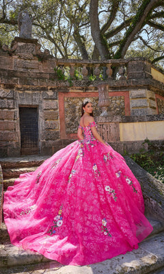 A young woman wears the Quinceanera Dress 54200 By Amarra, a bright pink off-the-shoulder gown with floral embroidery, as she stands on stone steps before an old stone structure among trees.