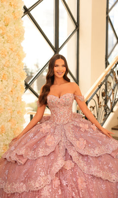 A woman smiles at the camera, standing indoors near a floral wall and staircase, wearing Quinceanera Dress 54202 By Amarra—an off-the-shoulder ball gown with lace embroidered flowers.