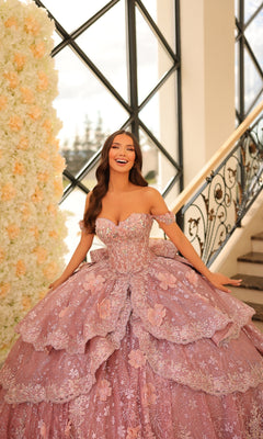 A woman in the Quinceanera Dress 54202 By Amarra, a pink lace ball gown embroidered with flowers, smiles near a staircase beside a wall adorned with white blooms.