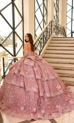 A woman in the Quinceanera Dress 54202 By Amarra, an elegant off-the-shoulder ball gown with lace embroidered flowers, stands by a staircase in a bright room with large windows.