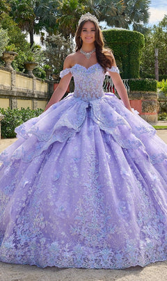 A young woman stands outdoors in a garden wearing the Quinceanera Dress 54202 by Amarra, an off-the-shoulder lavender ball gown with lace embroidered flowers, paired with a tiara and jeweled necklace.