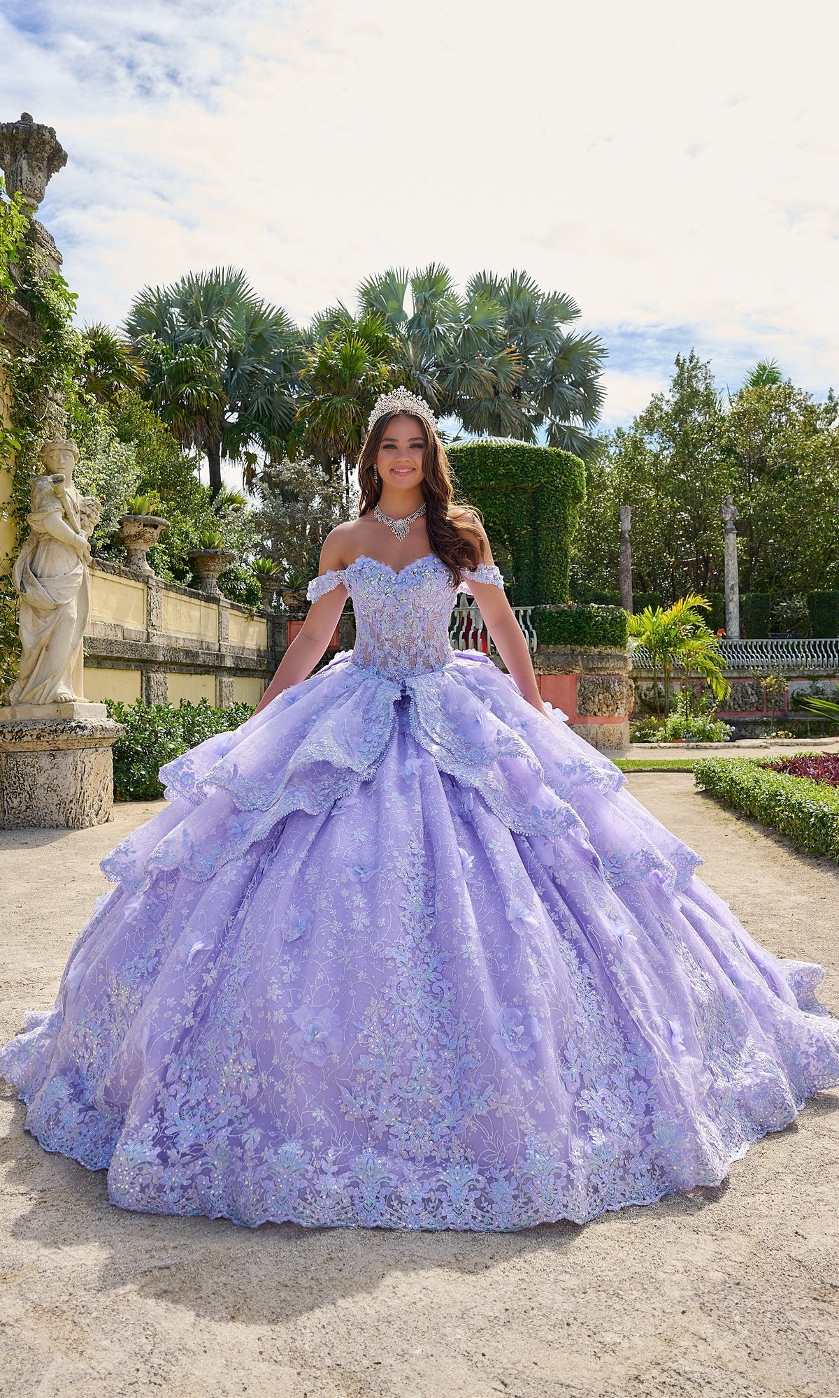 A young woman stands in a garden wearing the Quinceanera Dress 54202 by Amarra, a lavender ball gown with a full skirt, off-the-shoulder design, lace embroidered flowers, and a tiara.