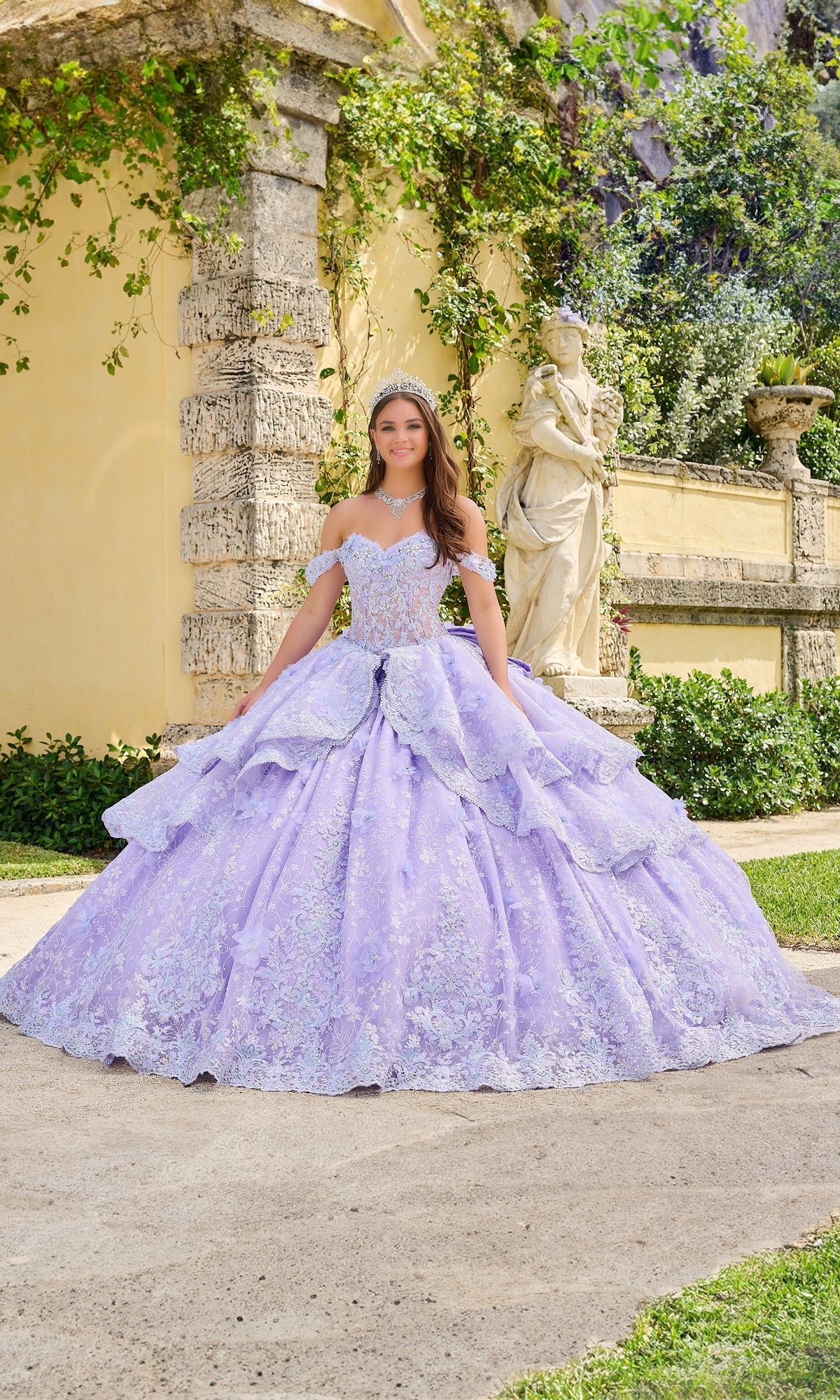A young woman stands outdoors by stone walls and greenery, wearing the Quinceanera Dress 54202 By Amarra—an off-the-shoulder lavender ball gown with lace embroidered flowers and a full skirt.