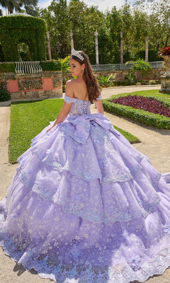 A young woman wearing the Amarra Quinceanera Dress 54202, an off-the-shoulder lavender ball gown with a large bow and tiara, stands in a formal garden with her back to the camera.