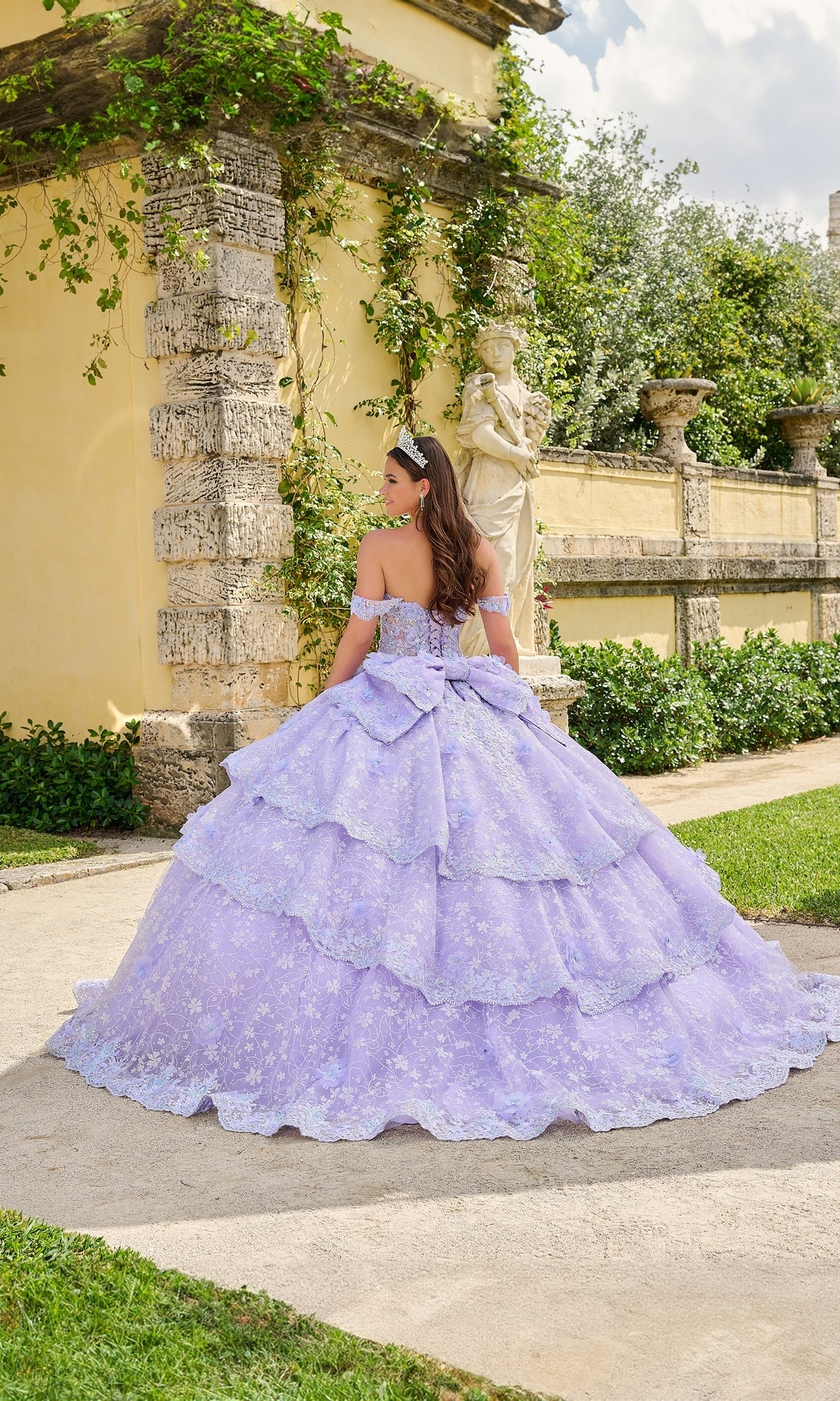 A young woman faces away from the camera near a stone wall and statue, wearing the Quinceanera Dress 54202 By Amarra—an off-the-shoulder lavender ball gown with lace and ruffles.