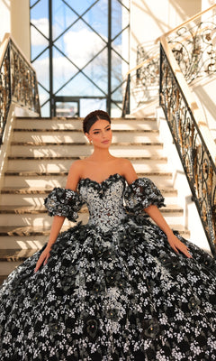 Wearing the Quinceanera Dress 54206 By Amarra, a woman in a strapless sweetheart ball gown with black and white floral details stands on a staircase with decorative railings and large windows behind her.