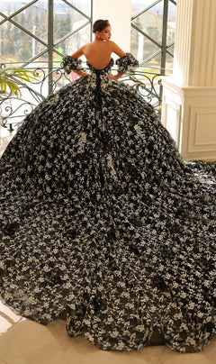 Indoors by a window, a person wears the Quinceanera Dress 54206 By Amarra—a dramatic off-the-shoulder ball gown with black and white floral patterns and a sweeping train.