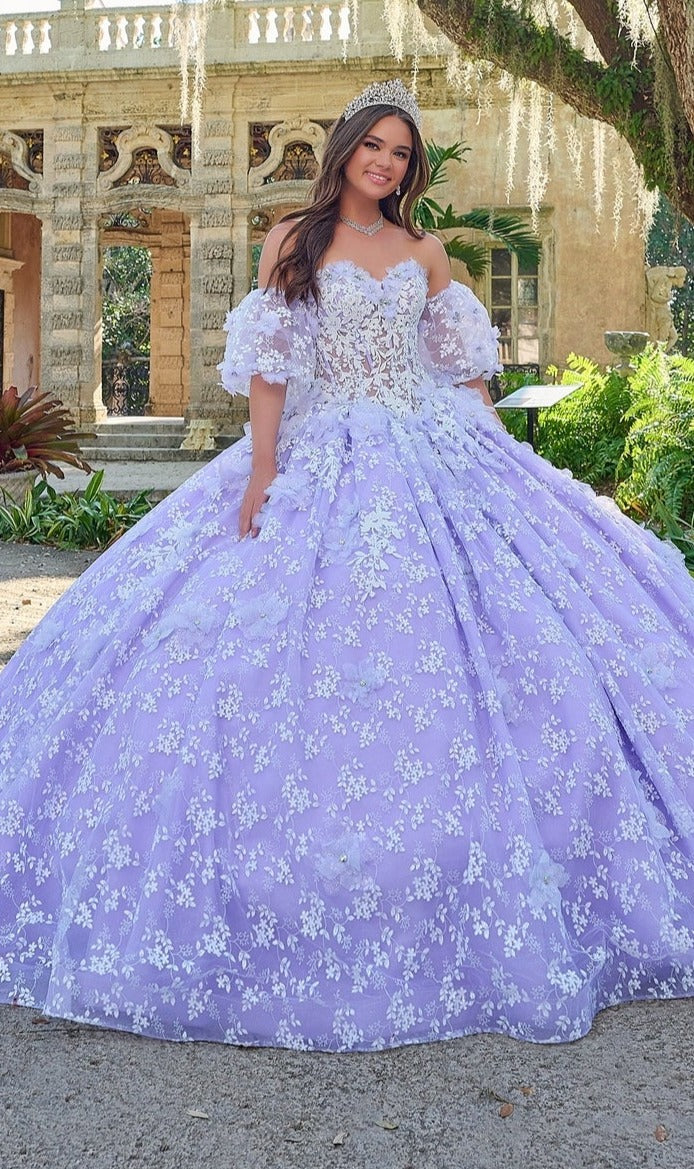 A young woman in the Quinceanera Dress 54206 By Amarra, a lavender ball gown with floral lace details, stands smiling outdoors in front of an ornate stone building.